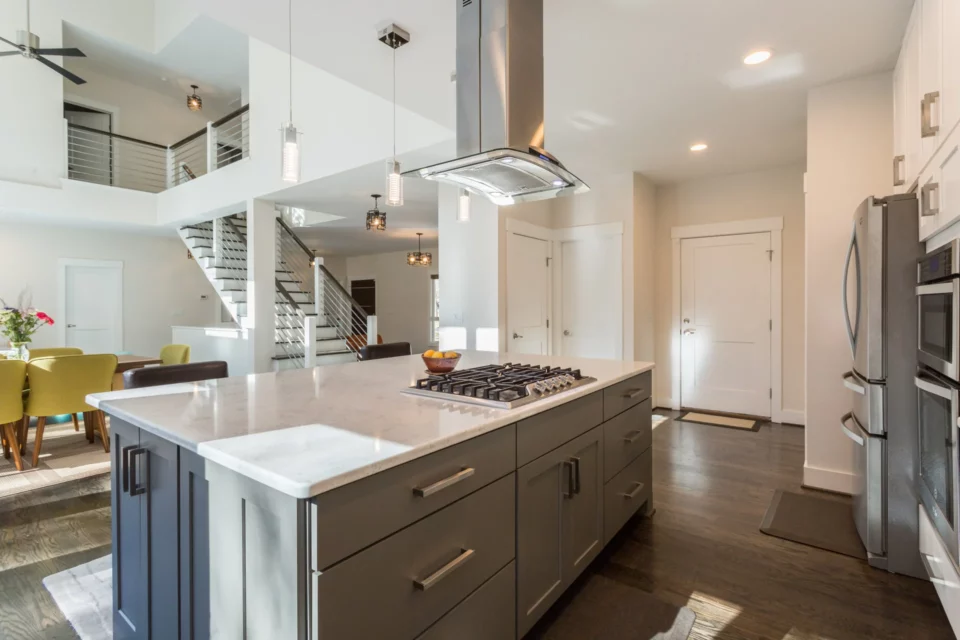 Kitchen island with gas cooktop, vent hood, and stainless refrigerator under a vaulted ceiling near the floating staircase