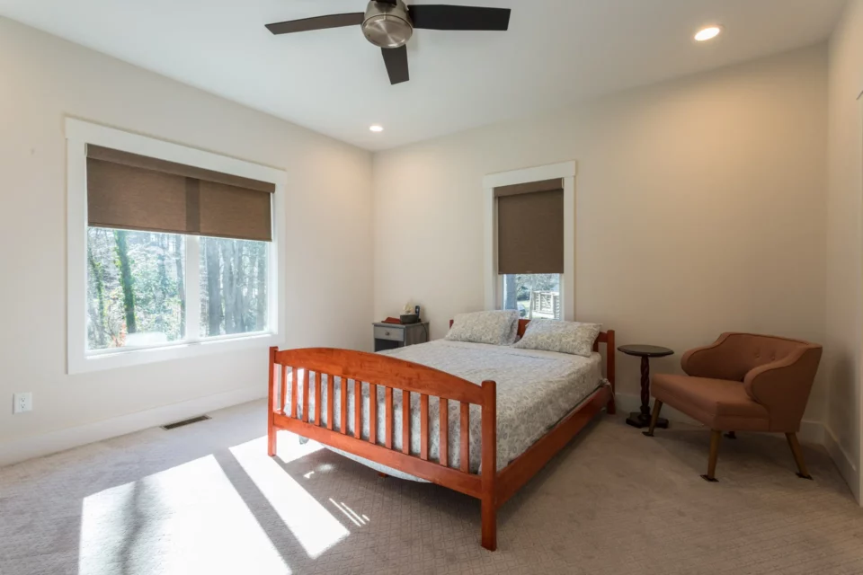 Carpeted bedroom with a wood bed frame, ceiling fan, neutral palette, and a sitting chair by the window