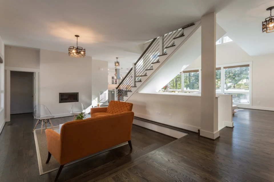 Living area with orange velvet chair and white seating beneath a vaulted ceiling, floating stairs with cable railings visible behind