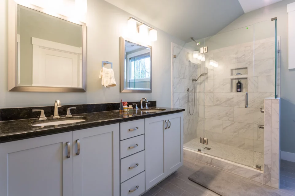 Primary bathroom with double vanity, dark granite counters, and a frameless glass walk-in shower with marble tile