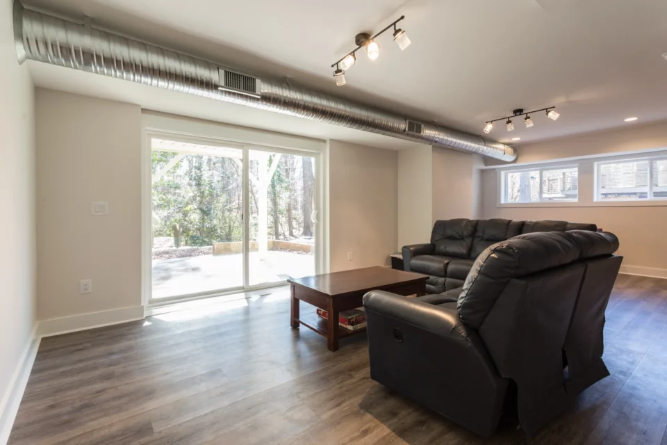 Finished basement family room with brown leather recliner sofa, exposed silver ductwork ceiling detail, and sliding glass door to the yard