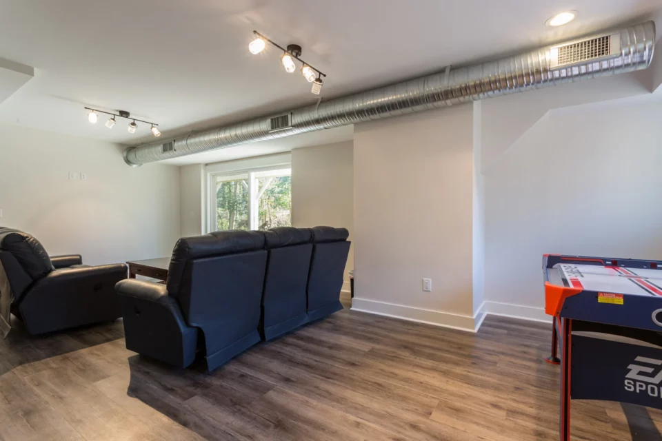 Finished basement game room with dark sectional, arcade machine, and track lighting under exposed ductwork